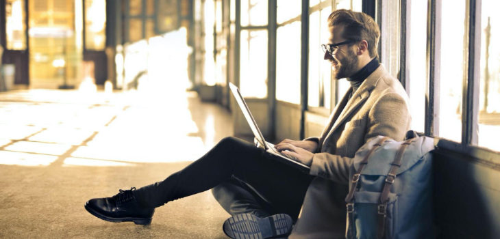business casual person sits on floor using laptop and smiling